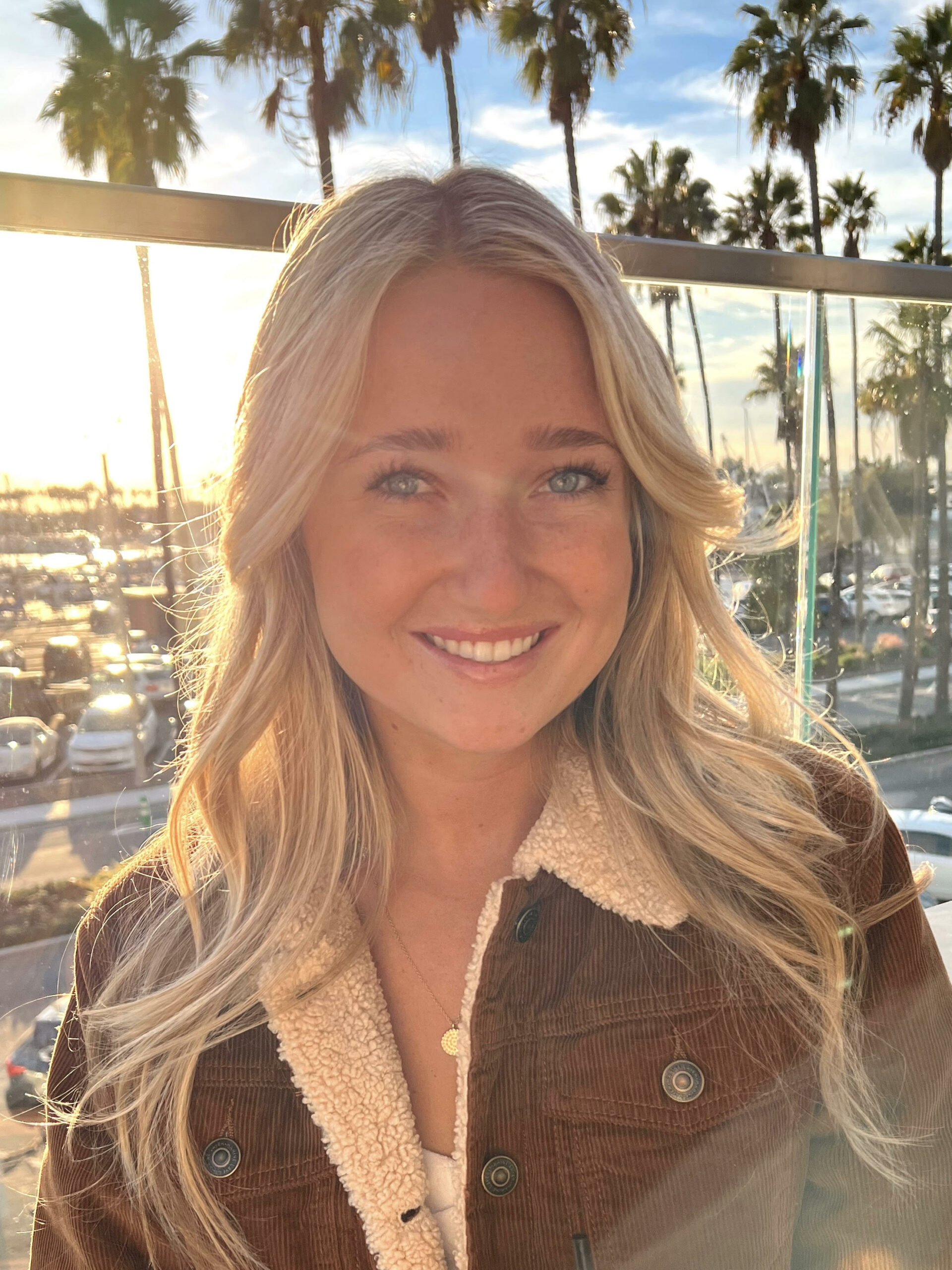 Blonde woman in brown corduroy jacket smiles outdoors near palm trees.